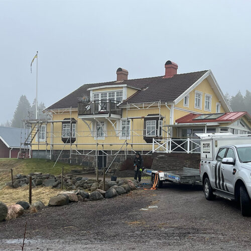 Gult trähus med byggställningar runt om under renovering, dimmig dag med arbetsfordon framför
