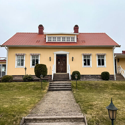 Gult traditionellt hus med rött tegeltak och symmetrisk fasad, fotograferat framifrån med stenlagd trappa och välskött trädgård.