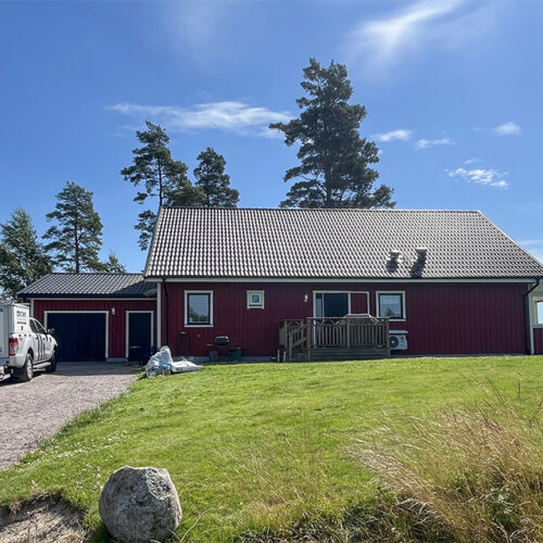 Rött enplanshus med svart tak på landet, byggfordon och material syns framför huset, vilket antyder pågående bygg- eller renoveringsarbete.