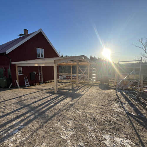 Pågående byggnation av träkonstruktion vid lantligt hus i solnedgång, med ställningar och byggmaterial på plats.
